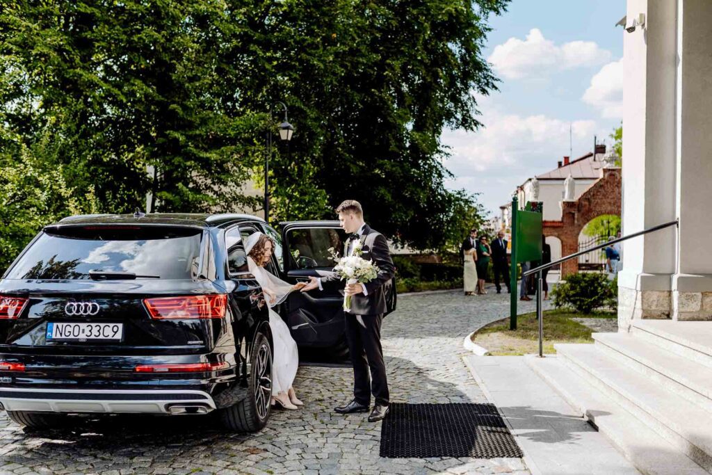 Pan Młody pomaga Pannie Młodej wysiąść z czarnego Audi przed kościołem.
