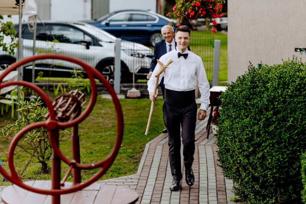 Uśmiechnięty Pan Młody idący chodnikiem przed domem, trzymający w dłoni drewnianą laskę.